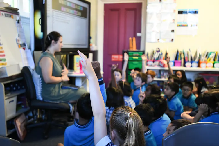 A primary teacher speaks to students, sitting on the floor.