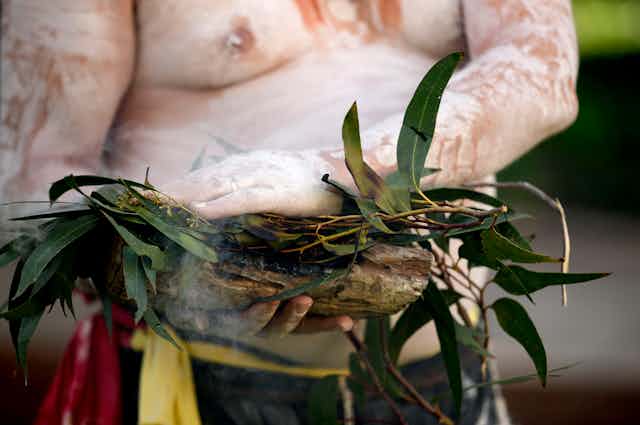 A man painted in white ochre performs a smoking ceremony during NAIDOC Week. He uses gum leaves in a coolamon.