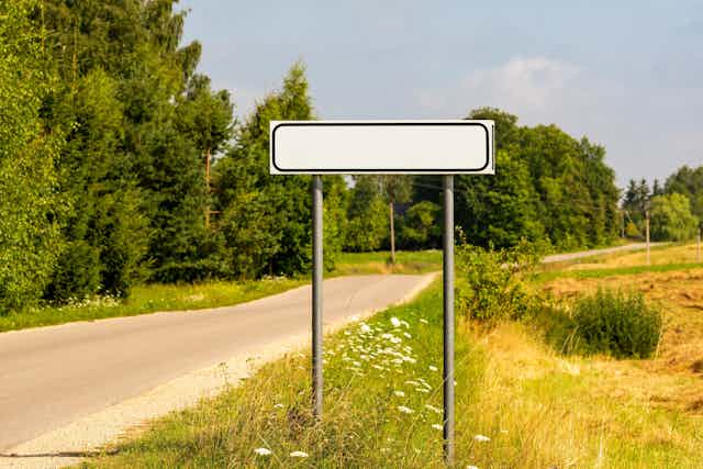 Un cartel de topónimos en blanco en medio de una carretera local.