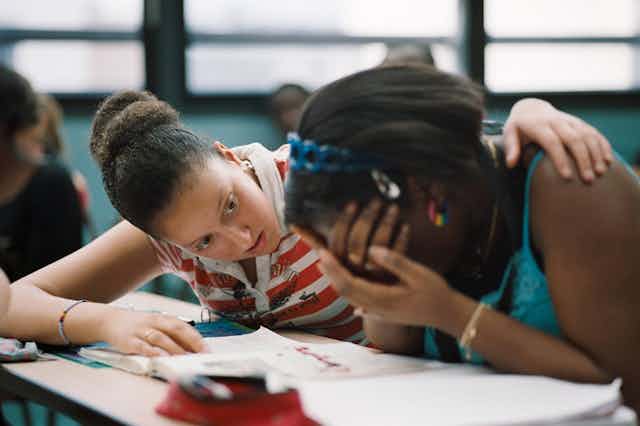 A high-school student hugs another student who cries on her desk.