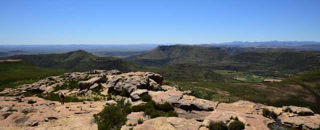 A panoramic photo of a mountain range, with a rocky outcrop in the foreground and a man standing on it.
