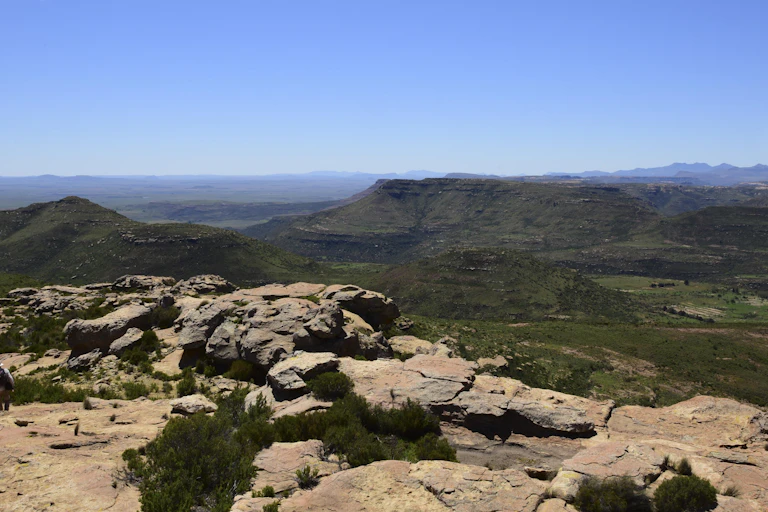 A panoramic photo of a mountain range, with a rocky outcrop in the foreground and a man standing on it.