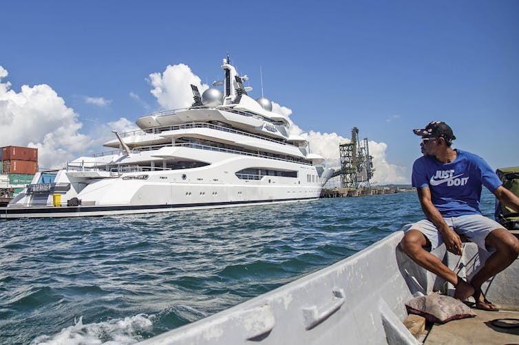 A man in a blue Nike t-shirt that says just do it look across a narrow channel to a massive yacht.