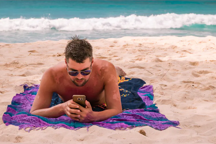Un homme regarde son téléphone sur la plage, le dos et les bras couverts de cicatrices après avoir pelés
