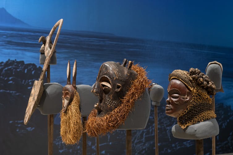 Displayed African masks and objects, two showing a male and female faces, seen against a blue landscape background.
