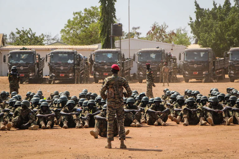 A man in military fatigues and a red cap stands in front of a group of men wearing military uniform and seated on dusty ground. Six parked military truck are behind them