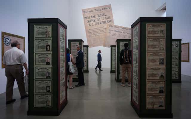 People walk around a museum exhibit. A large newspaper printour is displayed with the headline: must bar oriental completely to save BC for white race.
