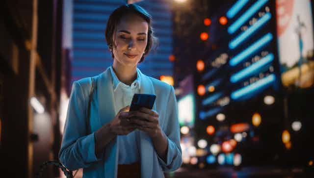 A young woman looking confidently at her mobile phone while standing on a city street at night