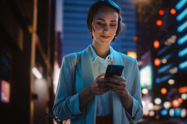 A young woman looking confidently at her mobile phone while standing on a city street at night