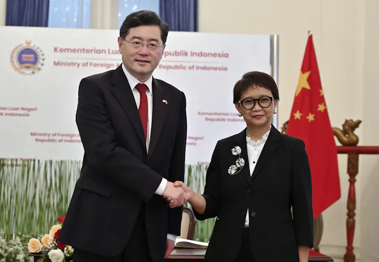 A short dark-haired woman shakes hands with a man in a dark suit and glasses.
