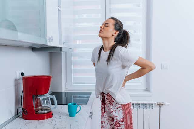 A young woman, standing in the kitchen near her coffeemaker, braces her lower back with her hands as she grimaces in pain.