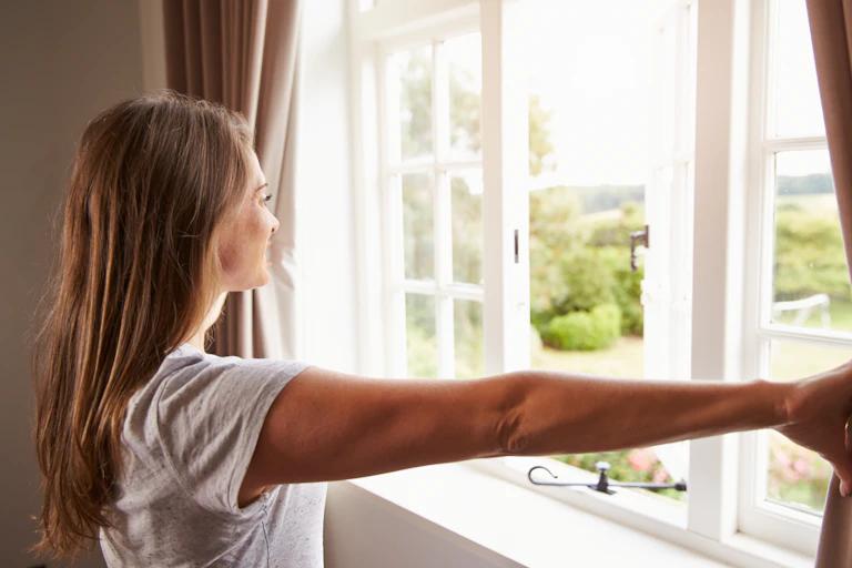 A woman holds back curtains in front of an open window