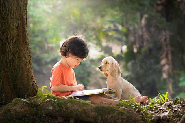A child reading under a tree next to a puppy.