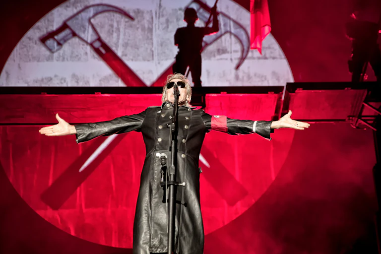 A man in a long black leather trench coat and sunglasses singing on stage.