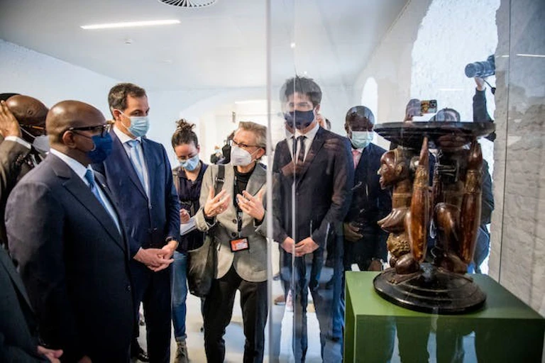 Des hommes et des femmes portant des masques se tiennent devant une sculpture en bois représentant deux personnages, l'un aux seins nus, avec des plateaux concaves en haut et en bas. Elle se trouve à l'intérieur d'une vitrine en verre