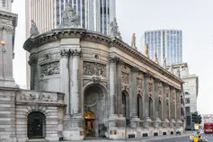 An ornate stone building with arched door and window, topped by statues. Skyscrapers in the background.