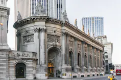 An ornate stone building with arched door and window, topped by statues. Skyscrapers in the background.