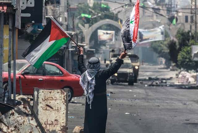 A man in an Arabic headscarf waves a Palestinian and a Syrian flag amid rubble and with military vehicles oncoming. 