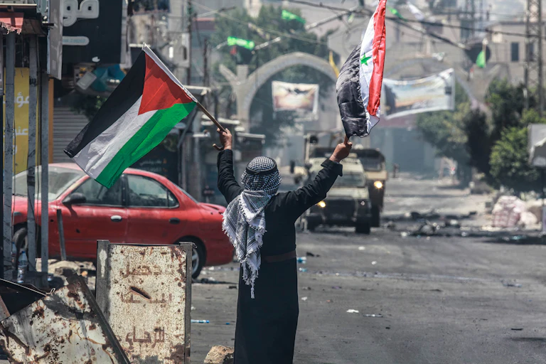 A man in an Arabic headscarf waves a Palestinian and a Syrian flag amid rubble and with military vehicles oncoming.