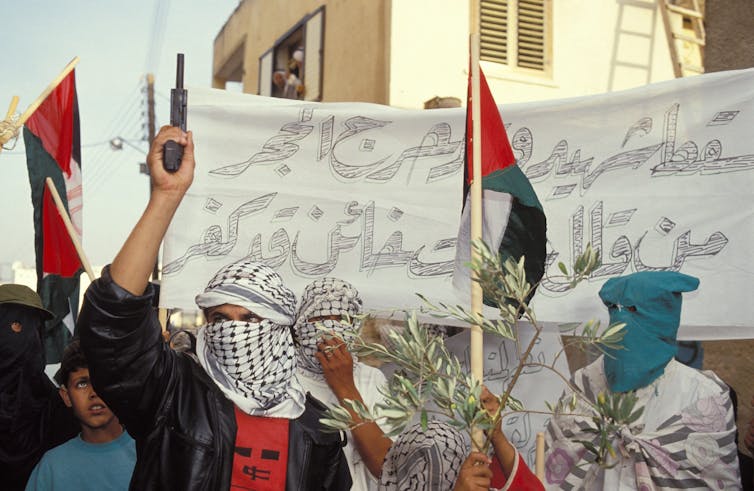 A group of men in headscarves stand in front of flags and banners. One holds a pistol up in the air.