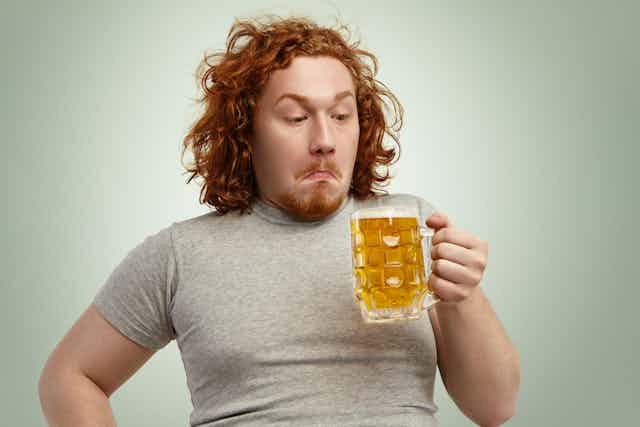 Red-haired young man with grey t-shirt and curly hair holding glass of light beer, looking confused or indecisive, standing against blank wall.