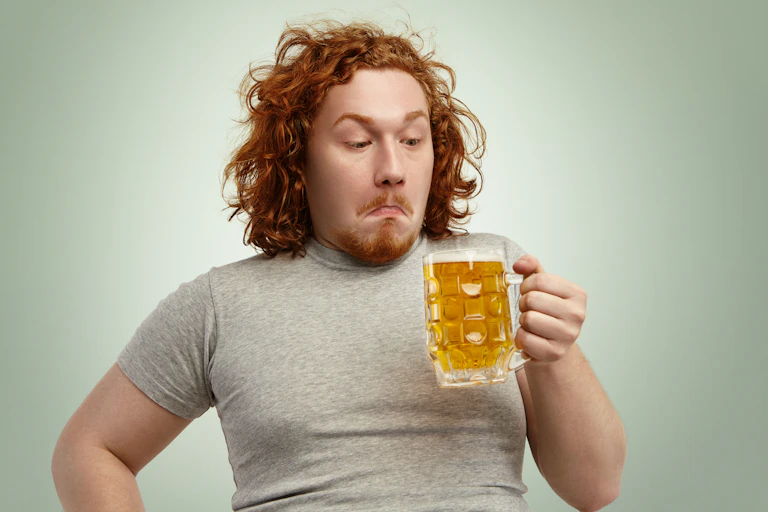 Red-haired young man with grey t-shirt and curly hair holding glass of light beer, looking confused or indecisive, standing against blank wall.