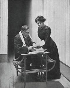 Un hombre sentado en una mesa con abrigo y bufanda habla con una mujer, de pie a su lado.