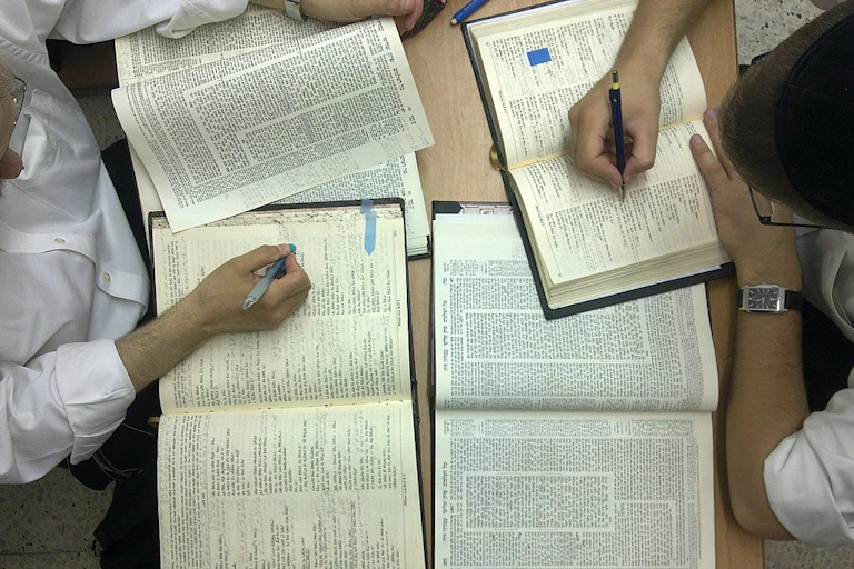 Two hands holding pens hover over open religious texts with writing densely covering the pages.
