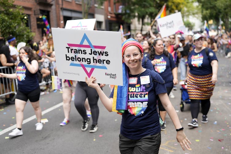 People in t-shirts that include the phrase 'queer pride' walk in a parade, with one holding a sign that says 'Trans Jews belong here.'