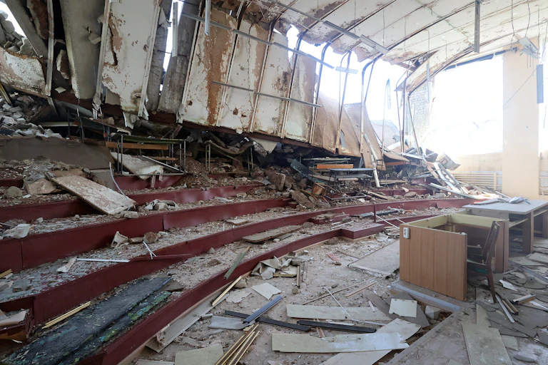 A destroyed classroom with a desk in the front of bleacher seats.