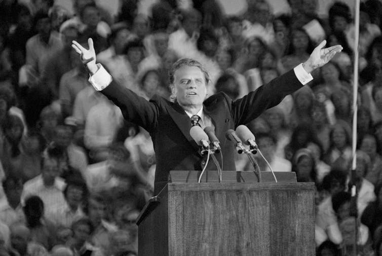 Black and white photo of Billy Graham preaching to a packed audience. Graham stands at a lectern in front of many onlookers, with his hands raised above his head