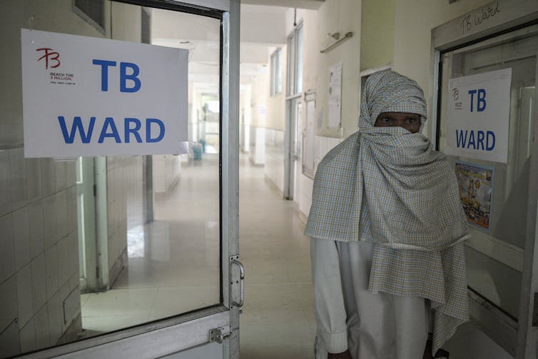 An man standing in a hospital next to a sign with TB Ward written on it.