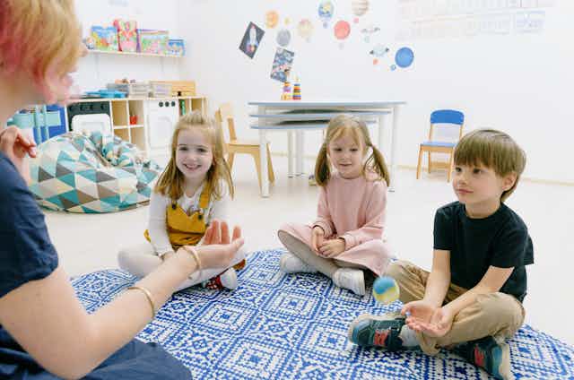 A childcare educator sits with three children on a mat.