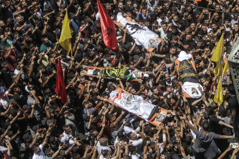 Stretchers carrying four dead people are carried by a crowd.