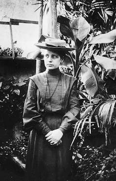 A young woman with her hands clasped together standing in front of a large plant and wearing a skirt, blouse and hat.