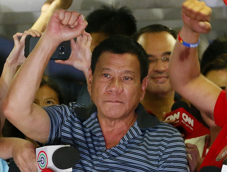 A man in a blue striped shirt, standing among a number of people, raises his clenched fist.