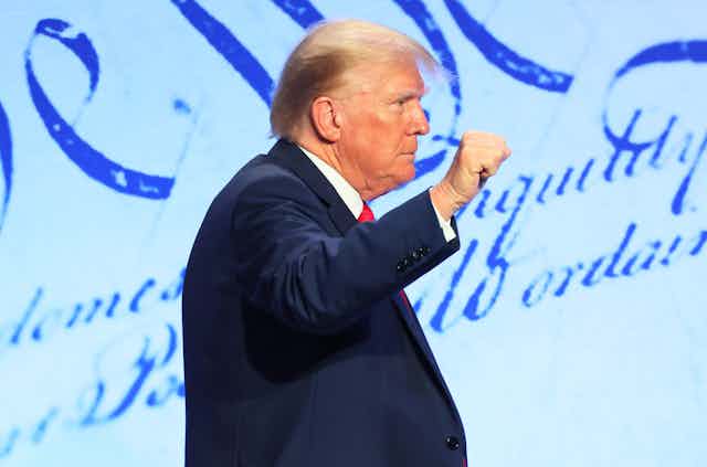 A man in a blue suit jacket, white shirt and red tie with his right fist raised.