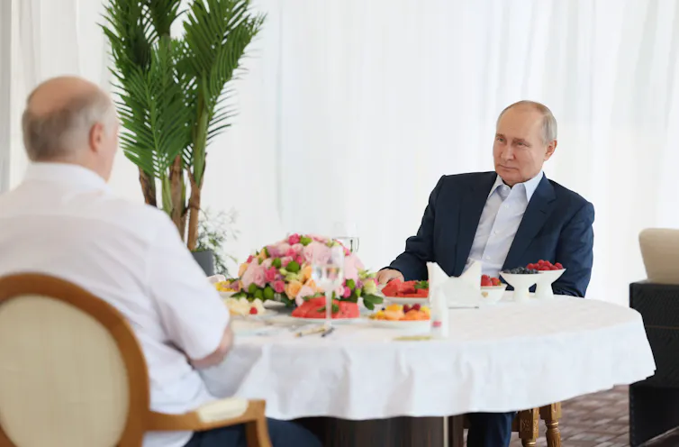 Putin sits at a table laid with food opposite another man.