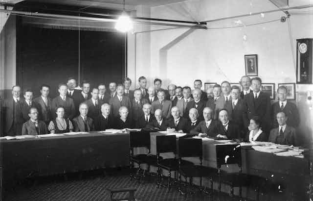 About thirty people stand in a conference room in a black and white photo. They are mostly white men wearing suits, with two women in the front row. 