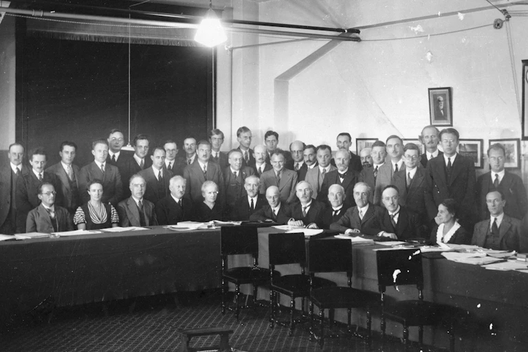 About thirty people stand in a conference room in a black and white photo. They are mostly white men wearing suits, with two women in the front row.
