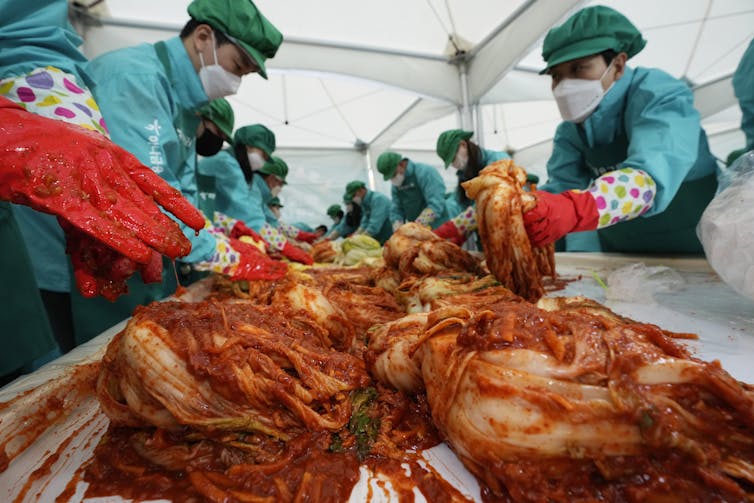 People in scrubs wearing masks and gloves handle mounds of fermented cabbage with red chilis colouring them red.