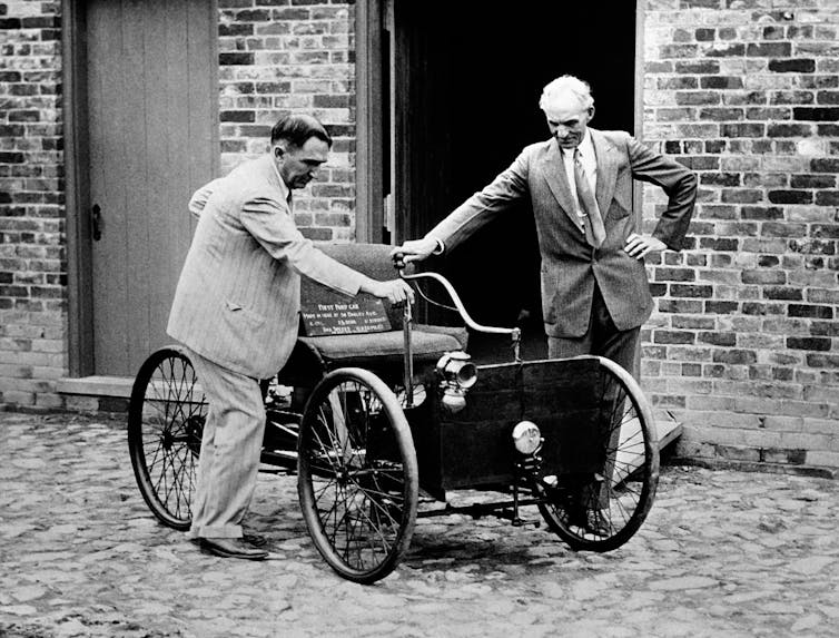 Two men with hands on an old model of a car from the 1890s.