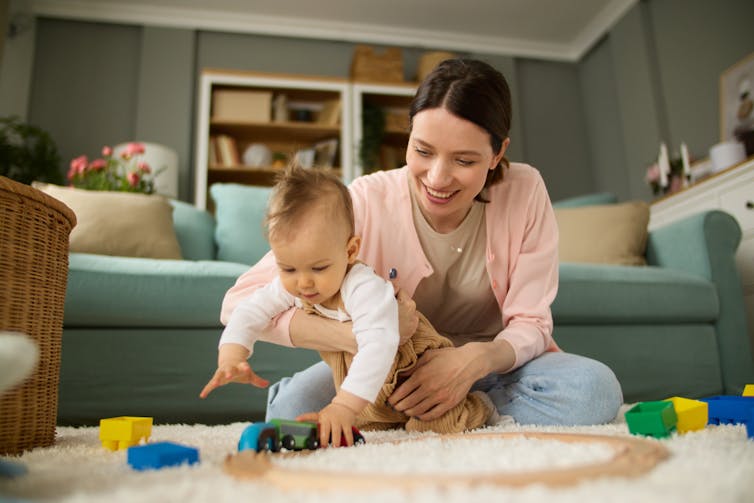 A woman on the floor at home with her baby who is playing with toys.