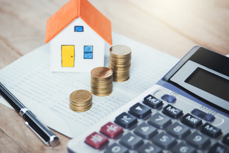 A pile of coins sits next to a calculator and a small house figurine.