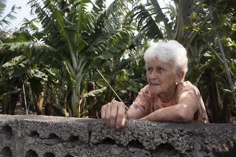A mature woman leans against a stone wall with banana trees in the background.