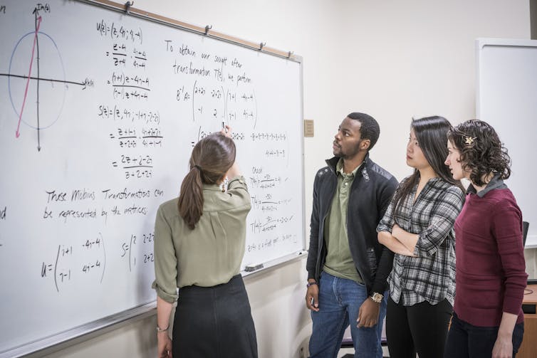 Three students (two women and one man) watch a woman professor write equations on a whiteboard.