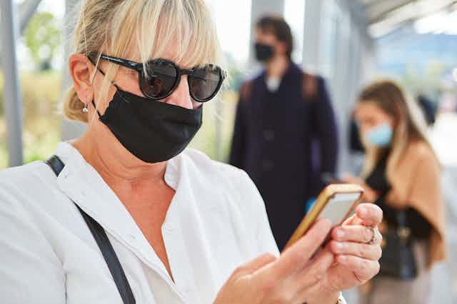a woman wearing sunglasses and a mask looks at a smartphone