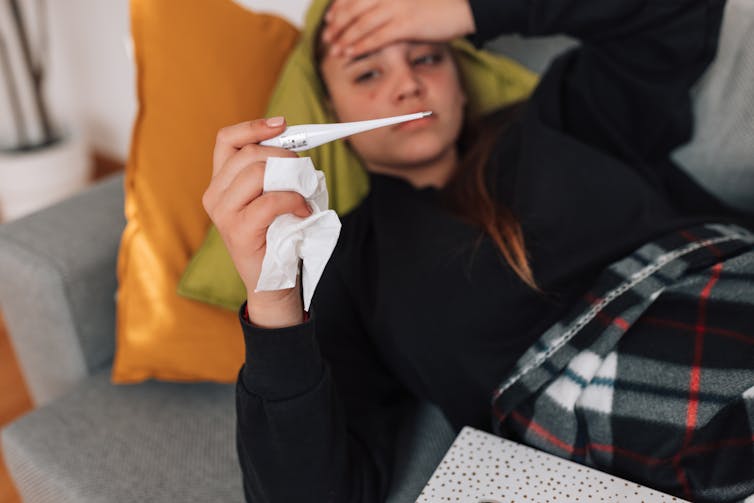A teenage girl is lying on a sofa, feeling unwell and holding a thermometer in her mouth to check her temperature.