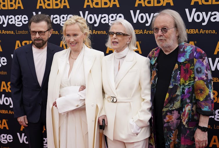 Four people, two men and two women, wearing fancy suits, stand at a red carpet event with the word ABBA behind them.