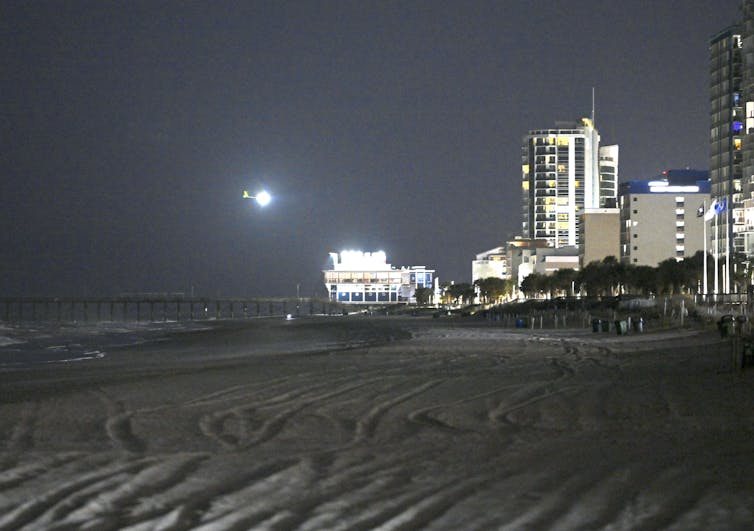 An illuminated spy balloon flies in the night sky near lit buildings and above a sanded section of a beach near the ocean.